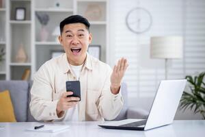 asombrado oriental hombre utilizando celular teléfono mientras sentado en remoto espacio de trabajo con personal computadora y cuaderno. extático adulto masculino bancario transacción pago en digital dispositivo durante jornada laboral. foto