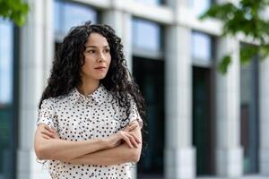 retrato de un pensativo Hispano mujer vistiendo un polca punto camisa, en pie con confianza al aire libre con cruzado brazos cerca un moderno edificio. foto