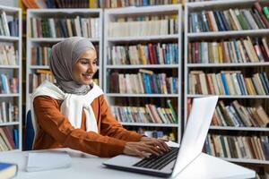 joven hermosa estudiante en hijab estudiando en línea remotamente en Universidad biblioteca, mujer mecanografía en ordenador portátil teclado buscando para información y preparando para examen. foto