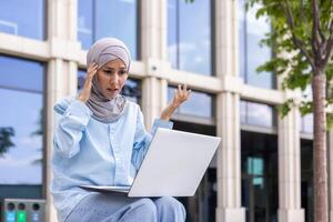 Young Muslim woman in a hijab looking confused while using a laptop on a university campus, showing frustration and technology challenges. photo