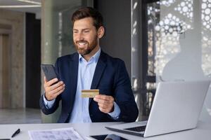exitoso financiero transacción y crecimiento. un sonriente y satisfecho joven empresario es sentado a el escritorio en el oficina, participación un banco crédito tarjeta y mirando a el teléfono pantalla. foto