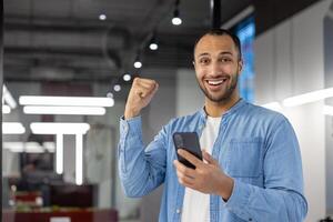 retrato de un contento musulmán hombre en pie en un moderno oficina, participación un teléfono en su mano y alegría en éxito y noticias, demostración un mano gesto, mirando a el cámara. foto