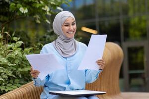 un alegre musulmán mujer en un hijab participación documentos, sonriente al aire libre con un borroso verde fondo, retratar confianza y positividad foto