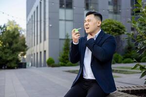 Suffering from a sore throat, a young Asian male businessman sits on a bench outside an office center and sprays medicine aerosol down his throat, holding his hand to his neck. photo