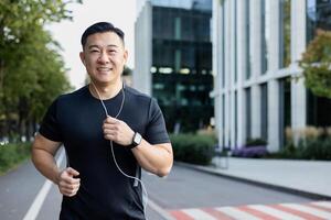 Portrait of an Asian young man running in headphones on a city street in the morning, smiling at the camera. photo