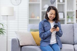 A young beautiful Asian sits alone at home on the sofa, the woman uses an application on the phone, holds a smartphone in her hands, smiles happily, uses the application, browses online pages. photo