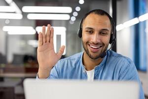 Close-up photo of a young smiling Arab man in a headset talking on a call on a laptop while waving his, hand at the camera.