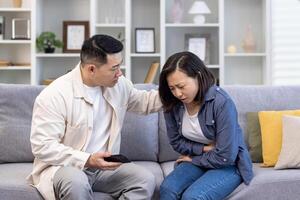 A young Asian man comforts and supports a woman sitting on the sofa at home and holding her stomach. Suffers from menstrual pain, pregnancy, miscarriage, poisoning. photo