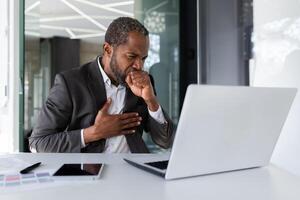Sick senior mature african american boss coughing at workplace inside office, businessman working with laptop sitting at desk. photo
