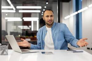 Disappointed and upset young hispanic man in blue shirt sitting at desk with laptop in office, looking displeased and waving hands at camera. photo