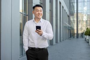 Smiling young Asian man walking near office center outside, holding phone in hand and looking away. photo