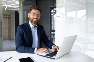 Portrait of a young businessman working on a laptop, sitting at a desk in the office and looking at the camera with a smile. photo