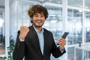 Portrait of happy hispanic businessman, man celebrating achievement good results and victory, boss looking at camera inside office holding phone, entrepreneur working inside office. photo