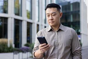 Close-up photo of a young Asian male businessman standing outside an office center with a phone in his hands, waiting for an appointment, making a call, typing a message.