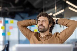 A content businessman reclines in his chair, eyes closed and smiling, enjoying a peaceful break in a lively office setting. Sticky notes in the background suggest a collaborative environment. photo