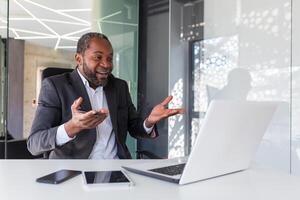 call online meeting mature african american businessman talking to colleagues remotely using laptop, man in business suit smiling gesturing cheerfully with hands inside office. photo