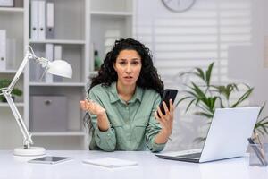 Perplexed mature Latina professional at desk with laptop and mobile phone, managing work tasks in a bright home office. photo