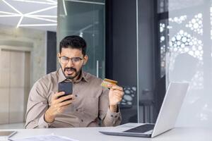 Indian businessman looking puzzled while holding a credit card and using his smartphone in a modern office setting. Concept of financial confusion or fraud. photo