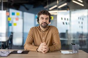 profesional hombre en un marrón camisa vistiendo un auriculares, sentado con confianza a su lugar de trabajo en un moderno oficina. foto