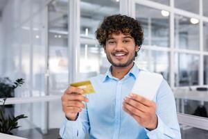 un joven Hispano hombre es sentado en el oficina, participación un teléfono y utilizando un crédito tarjeta. sonriente señalando a el cámara. foto