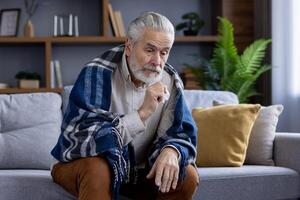 mayor hombre con gris pelo y barba envuelto en un acogedor frazada, mirando pensativo mientras sentado en un sofá en un elegante vivo habitación. foto