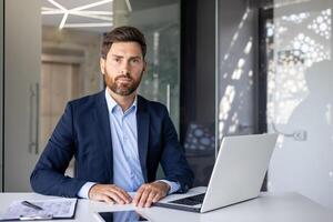 Portrait of a successful and confident young man lawyer in a business suit sitting at an office desk, working with a laptop and documents, looking seriously at the camera. photo