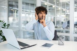 Young professional man in a blue shirt sitting at a white office desk, making a silence gesture with a finger to lips, surrounded by modern office environment. photo