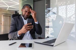 trastornado y decepcionado hombre dentro oficina participación teléfono, empresario leyendo malo Noticias a lugar de trabajo dentro oficina, africano americano mayor jefe infeliz con logro resultados notificación. foto