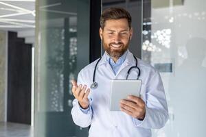 un joven sonriente masculino médico es en pie en el hospital vestíbulo en un blanco Saco y hablando vía comunicación con el paciente vía un tableta. él explica, consultas. foto