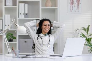 Joyful Latina doctor in white coat taking a break at her office desk with laptop, feeling content and happy. photo