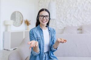 Young female blogger sitting on sofa at home in headset and chatting online on camera, smiles photo