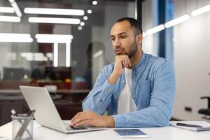 Pensive young hispanic man working and studying in office, sitting at desk holding head in hand and looking at laptop, analyzing data, looking for solution, thinking about project. photo