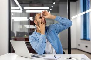 indio Hispano hombre en un vivo habitación configuración, trabajando en un oficina ambiente, utilizando medicinal ojo gotas mientras sentado a su escritorio. foto