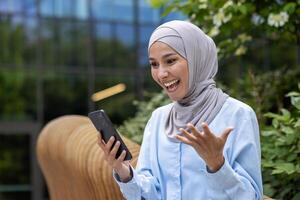 joven hermosa musulmán mujer en hijab usos teléfono fuera de oficina edificio, sonriente mujer lee en línea noticias, navega Internet paginas y diales mensaje llamar. foto