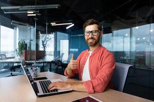 Portrait of joyful successful businessman at workplace, man smiling and looking at camera, showing thumbs up gesture, worker with laptop and glasses. photo