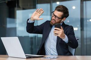 Young handsome man sitting in a suit in the office at the table and listening to music from the phone in headphones. Dances along, relaxed, waves his hands. photo