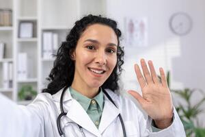 hembra médico sonriente y mirando a teléfono inteligente cámara ondulación mano alegremente saludo, Hispano mujer en blanco médico Saco hablando utilizando aplicación en teléfono para llamar, en pie dentro clínica. foto