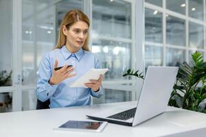 enfocado mujer de negocios en un azul camisa atractivo en un virtual reunión en un brillante oficina ajuste con un ordenador portátil y tableta. foto