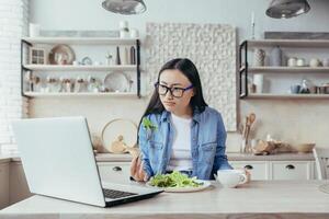 An interested Asian woman is sitting at the kitchen table at home. Carefully watches the online seminar on dietetics and healthy eating. He eats a fresh salad, holds a cup with a drink in his hand. photo