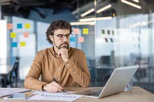 Concerned administrator resting head on clenched fist and looking at pc screen while making notes by hand. Attentive manager filling data from digital device and making remarks with main thesis. photo