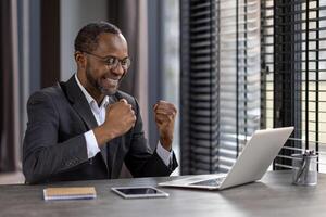 Joyful African American businessman with glasses expressing success while working on laptop in modern office. photo