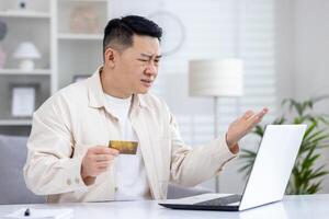 asiático hombre engañado por estafadores, hombre sentado a hogar a mesa participación banco crédito tarjeta, rechazado dinero transferir, negativo cuenta balance, trastornado y decepcionado. foto