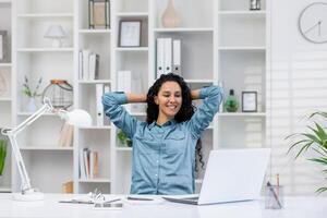 un relajado Hispano mujer toma un descanso mientras trabajando remotamente en su brillante hogar oficina, encarnando vida laboral balance. foto