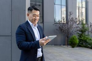 Smiling Asian male professional in a suit engaging with a tablet PC on a sunny day outside a corporate building. photo