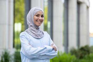 A professional Muslim woman in a hijab stands confidently outdoors with her arms crossed, exuding empowerment and elegance. photo