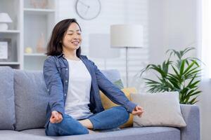 joven sonriente asiático mujer meditando sentado en sofá en vivo habitación, mujer sonriente alegremente con ojos cerrado, soñando y pensando acerca de futuro planes, visualizante logro. foto