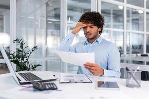 trastornado financiero mirando a ventas y ingresos gráfico de compañía, empresario dentro oficina decepcionado con ventas resultados y logros, Hispano hombre sentado a mesa con documentos y ordenador portátil. foto