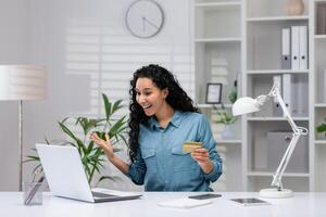 Joyful Hispanic woman with credit card making purchases online while working from home office, expressing excitement. photo