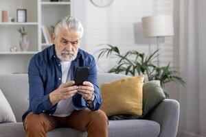 un maduro hombre con gris pelo intensamente enfoque en su teléfono inteligente en un moderno vivo habitación configuración, transporte un concepto de tecnología utilizar entre el anciano. foto