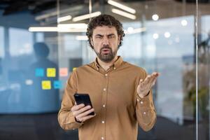 Confused casual male standing alone with smartphone in palm and looking at camera with irritation. Busy person getting notification about discharged battery of mobile gadget and feeling dissatisfied. photo